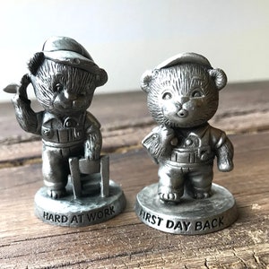 May include: Two pewter figurines of bears wearing hats and overalls. The bear on the left is holding a tool and the base reads "Hard at Work". The bear on the right is holding a tool and the base reads "First Day Back".
