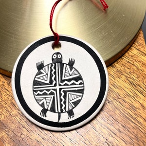 May include: A white ceramic ornament with a black and white geometric design of a turtle. The ornament has a red string attached to it.