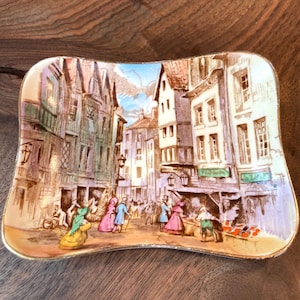 May include: A small, square, ceramic dish with a gold rim. The dish features a colorful painted scene of a European street with people walking and shopping.