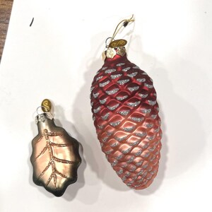 May include: Two glass Christmas ornaments. One is a large, red and copper-colored pinecone with silver glitter accents. The other is a smaller, leaf-shaped ornament with gold and copper tones. Both have gold hanging loops.
