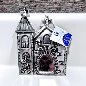 May include: A silver-colored metal church-shaped decorative piece with a cross on top. The piece has intricate cut-out designs and a small tag attached. The tag has the text "La Casa del Rebozo".