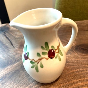 Puede incluir: Jarra de cer&aacute;mica blanca con un dise&ntilde;o floral verde y rojo. La jarra tiene un cuerpo redondeado y un asa.
