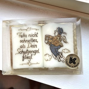 May include: A white candle shaped like an open book with a gold trim and the text "Fahr nicht schneller, als Dein Schutzengel fliegt."  The candle features a cartoon illustration of an angel with wings flying through the clouds. The candle has a gold seal with the text "Qualität in Wachs".