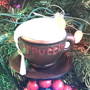 May include: A brown ceramic coffee cup ornament with white frosting and the word "Cappuccino" in red lettering. The ornament is hanging on a Christmas tree with red and green lights.