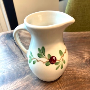 Puede incluir: Jarra de cer&aacute;mica blanca con un dise&ntilde;o floral verde y rojo. La jarra tiene un asa y un pico.