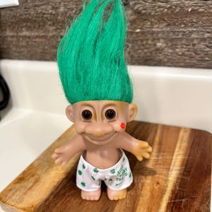 May include: A vintage troll doll with bright green hair, black eyes, and a small red kiss mark on its cheek. The doll is wearing white shorts with green shamrocks and the text "Irish Today". It is displayed on a wooden cutting board.