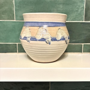 May include: A cream-colored ceramic pot with a rounded body and a flared rim. The exterior features horizontal blue bands and a repeating pattern of raised, textured, wave-like brushstrokes in a light tan color across the center section.