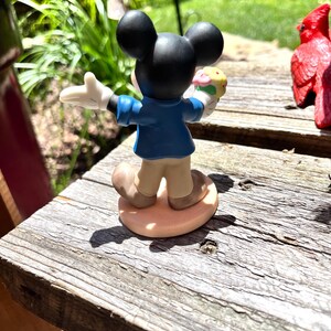 Puede incluir: Una figura de Mickey Mouse, vista desde atr&aacute;s, sosteniendo un helado colorido. Mickey lleva una chaqueta azul, pantalones color beige y zapatos marrones. La figura tiene orejas negras y guantes blancos. Est&aacute; sobre una base ovalada marr&oacute;n claro.