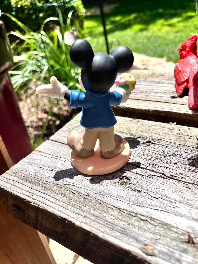 Puede incluir: Una figura de Mickey Mouse, vista desde atr&aacute;s, sosteniendo un helado colorido. Mickey lleva una chaqueta azul, pantalones color beige y zapatos marrones. La figura tiene orejas negras y guantes blancos. Est&aacute; sobre una base ovalada marr&oacute;n claro.