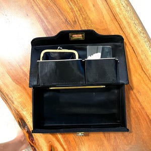 May include: Open black satin clutch with gold-tone hardware. The interior features a small coin purse, a pocket, and a mirror. The clutch is open, revealing the interior compartments. The clutch is on a wooden surface.