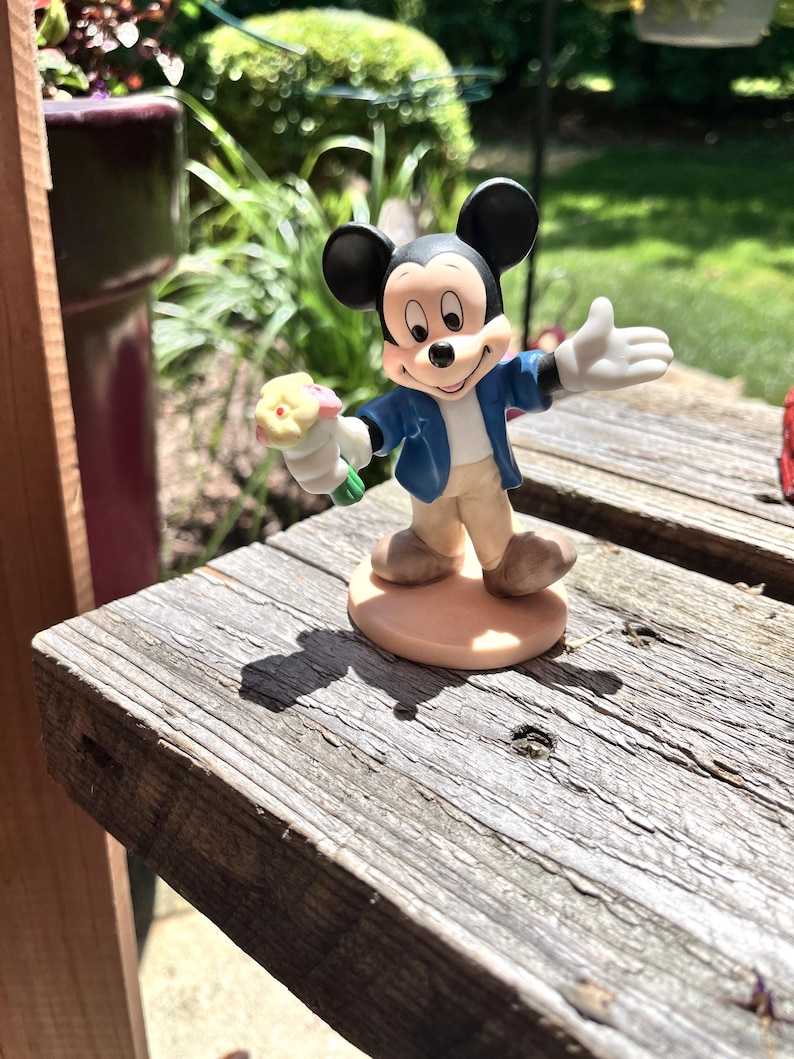 May include: A Mickey Mouse figurine stands on a weathered wooden surface. Mickey is depicted in his classic attire: a blue jacket, white gloves, and tan pants. He holds a small bouquet of flowers. The figurine is set against a blurred background of greenery and sunlight.