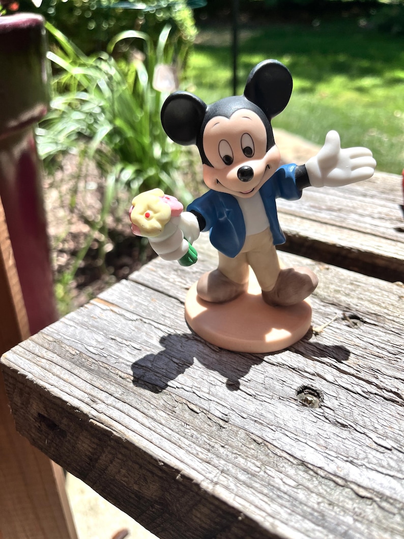 Puede incluir: Una figura de Mickey Mouse, con una chaqueta azul y pantalones color canela, sostiene un ramo de flores. La figura tiene orejas negras y zapatos marrones. La figura est&aacute; sobre una base redonda de color marr&oacute;n claro.