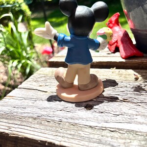Puede incluir: Una figura de Mickey Mouse, vista desde atr&aacute;s, sosteniendo una magdalena. Mickey lleva una chaqueta azul, pantalones color beige y zapatos marrones. La figura est&aacute; sobre una superficie de madera, con una estatua de cardenal rojo y vegetaci&oacute;n en el fondo.