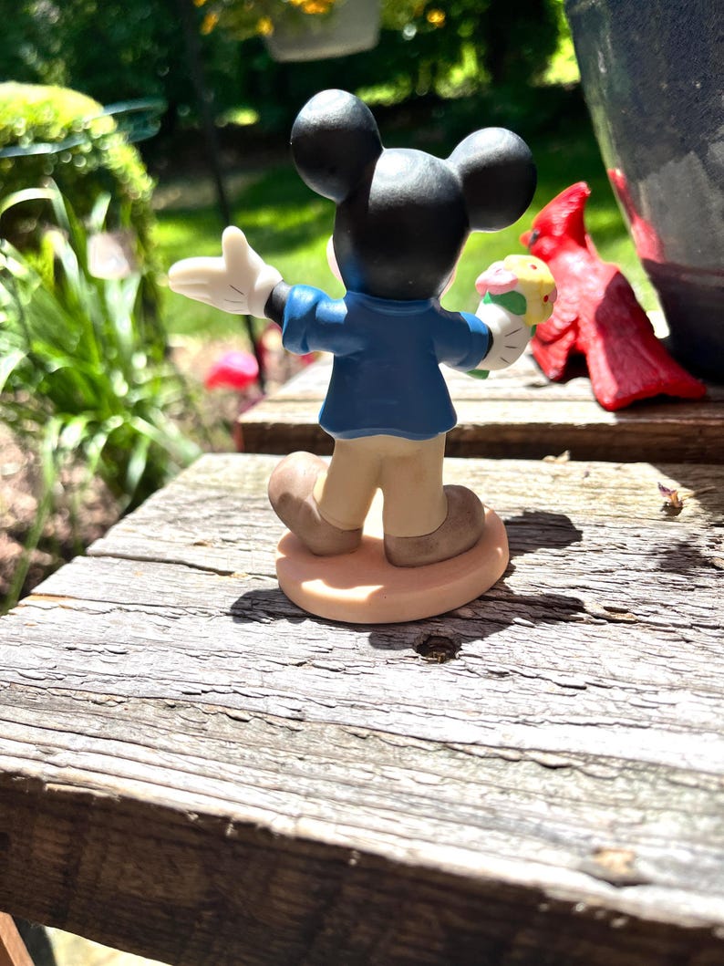 Puede incluir: Una figura de Mickey Mouse, vista desde atr&aacute;s, sosteniendo una magdalena. Mickey lleva una chaqueta azul, pantalones color beige y zapatos marrones. La figura est&aacute; sobre una superficie de madera, con una estatua de cardenal rojo y vegetaci&oacute;n en el fondo.