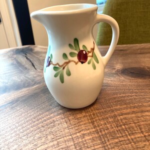 Puede incluir: Jarra de cer&aacute;mica blanca con un dise&ntilde;o floral verde y marr&oacute;n. La jarra tiene un cuerpo redondeado y un asa.