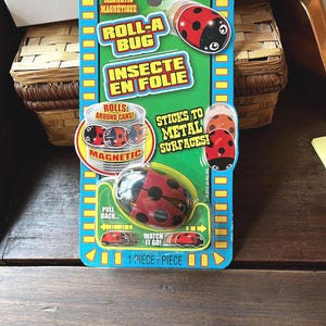 May include: A Roll-A-Bug toy package. The package is blue and green, with the words "Roll-A Bug" and "Insecte en Folie" printed on it. The toy is a ladybug that rolls and sticks to metal surfaces. The package also says "Rolls around cans!" and "Watch it go!"