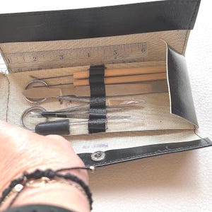 May include: A black faux leather folding case containing a set of metal and wooden tools, including two wooden-handled picks, a flat metal spatula, tweezers, and a dropper. A metal ruler is visible inside, measuring up to 6 inches in length.