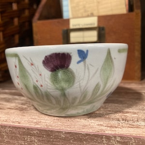 May include: A white ceramic bowl with a floral design featuring a purple thistle, green leaves, and blue flowers. The bowl is sitting on a wooden shelf.