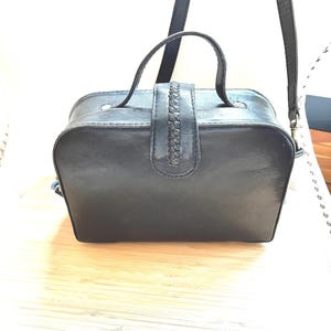 May include: Black leather handbag with a top handle and a braided strap closure. The bag has a rectangular shape with rounded edges and a detachable shoulder strap. The bag is sitting on a light wood surface.