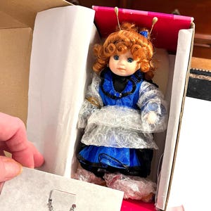 May include: A porcelain doll with red curly hair, wearing a blue and black dress with a white lace overlay. The doll is in a box with pink and white lining.