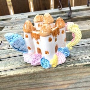 May include: A ceramic teapot shaped like a castle with a seahorse handle. The castle is decorated with brown, white, and yellow details. The base of the teapot is decorated with pink, blue, and yellow seashells.