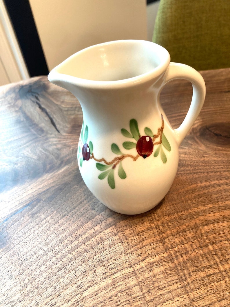 Puede incluir: Jarra de cer&aacute;mica blanca con un dise&ntilde;o floral verde y rojo. La jarra tiene un cuerpo redondeado y un asa.