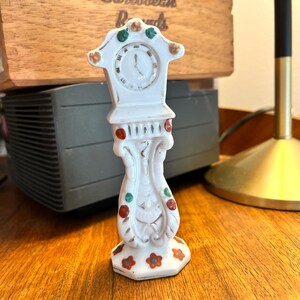 May include: White porcelain grandfather clock figurine with red, green, and gold accents. The clock face has black hands and Roman numerals.