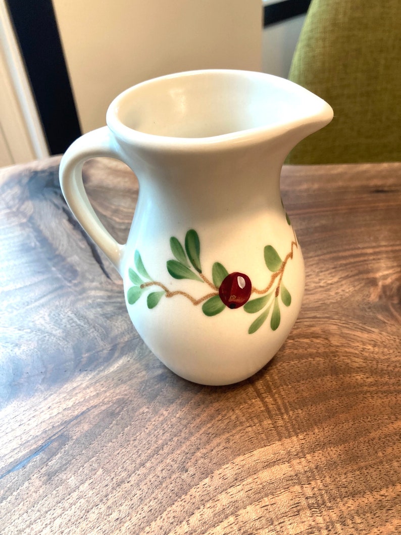 Puede incluir: Jarra de cer&aacute;mica blanca con un dise&ntilde;o floral verde y rojo. La jarra tiene un asa y un pico.