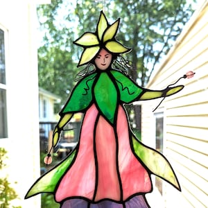 May include: A stained glass fairy figurine with a pink dress, green leaves, and a yellow flower crown. The fairy is holding a beaded wand.