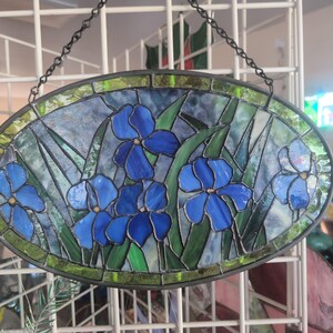 May include: Oval stained glass panel featuring blue iris flowers with green leaves against a blue and clear background. The panel is framed with a dark metal border and hangs from a chain. A decorative floral art piece.