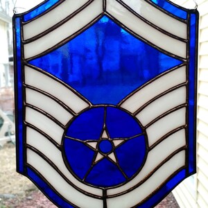 May include: A stained glass window depicting the rank insignia of a Technical Sergeant in the United States Air Force. The insignia features a blue background with a white five-pointed star in the center, surrounded by four white chevrons.