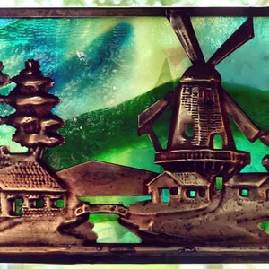 May include: A stained glass window depicting a windmill, houses, and trees. The scene is set against a green and blue background. The windmill is made of metal and has a brown finish. The houses are also made of metal and have a brown finish. The trees are made of metal and have a green finish.