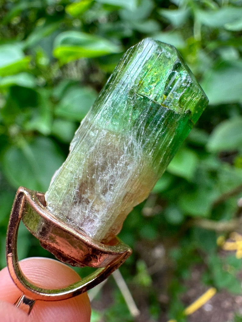 May include: A green and white crystal pendant with a gold setting. The crystal is a long, thin, and pointed shape with a striated surface.