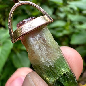 May include: A green, white, and pink tourmaline crystal pendant with a gold-tone setting. The pendant is attached to a gold-tone chain.