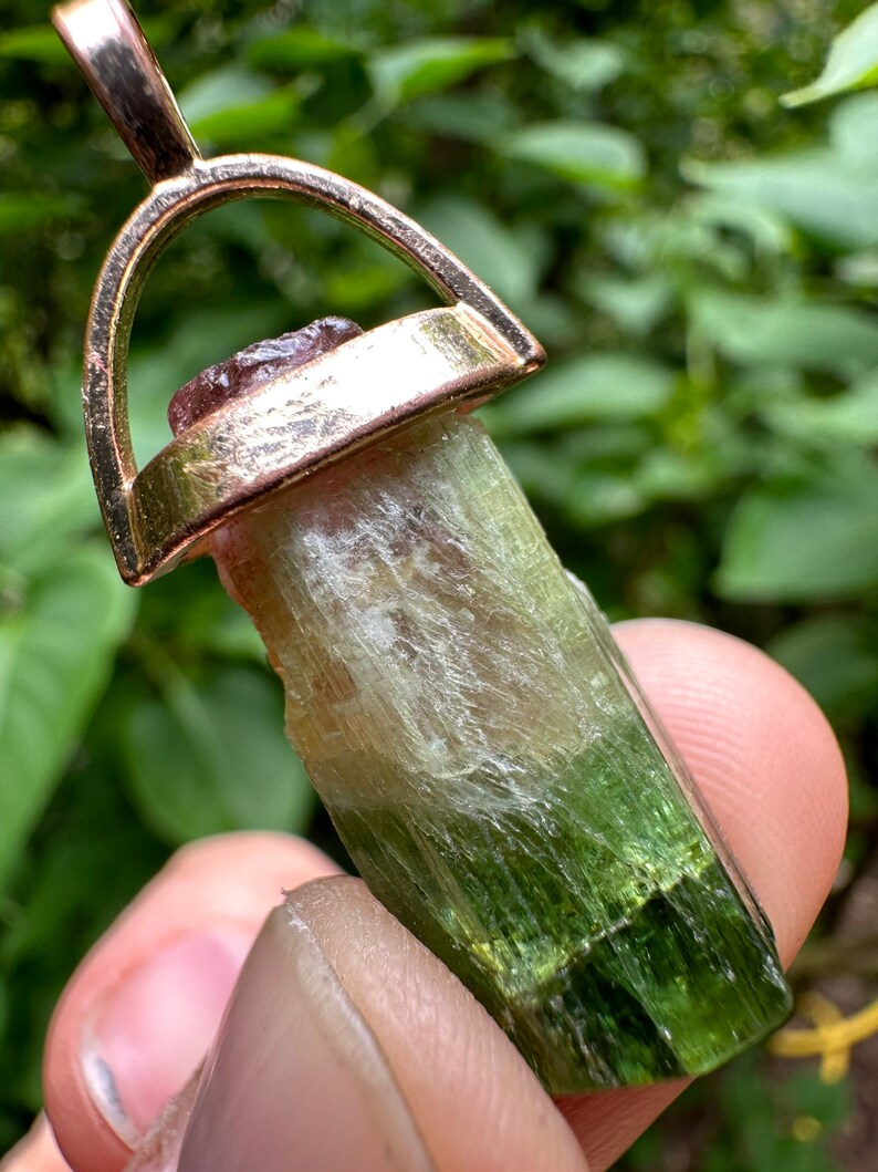 May include: A green, white, and pink tourmaline crystal pendant with a gold-tone setting. The pendant is attached to a gold-tone chain.