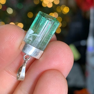 May include: A green tourmaline pendant with a silver setting. The pendant is a rectangular prism with striations on the surface. The pendant is hanging from a silver chain.