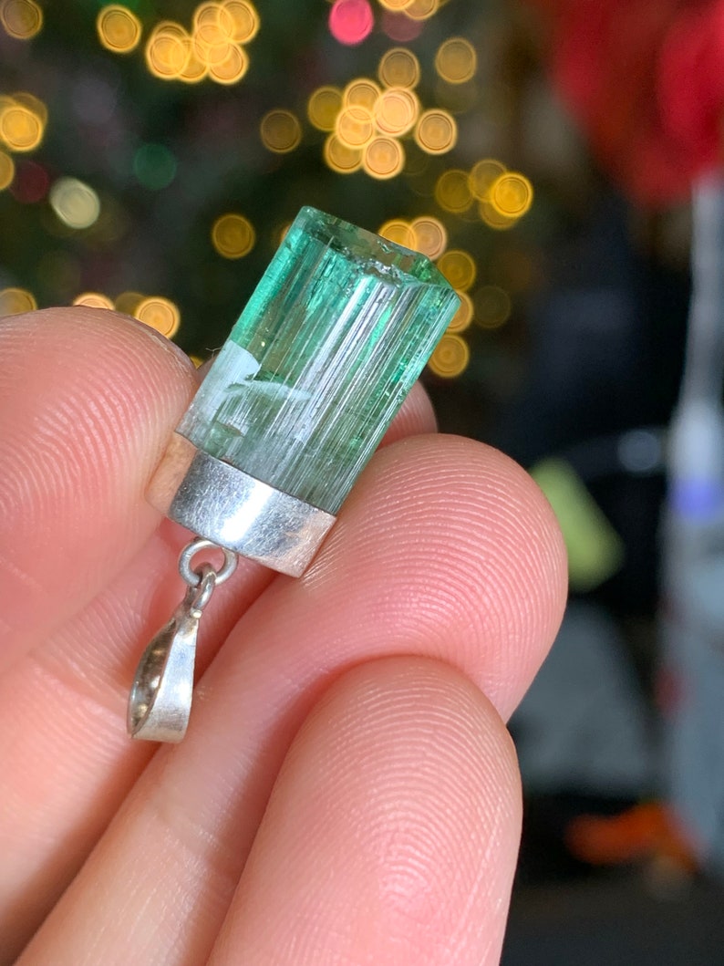 May include: A green tourmaline pendant with a silver setting. The pendant is a rectangular prism with striations on the surface. The pendant is hanging from a silver chain.