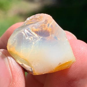 May include: A raw, translucent opal gemstone with a milky white and pale blue body color, featuring internal flashes of orange and yellow light. The stone has an irregular, multifaceted shape with smooth, polished surfaces and natural edges.