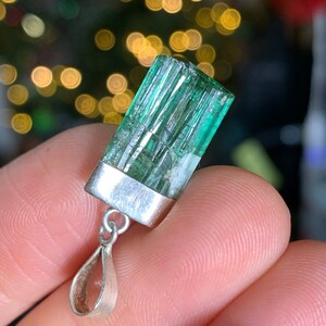 May include: A green gemstone pendant with a silver setting. The gemstone is a rectangular shape with a striated texture. The pendant is attached to a silver bail.