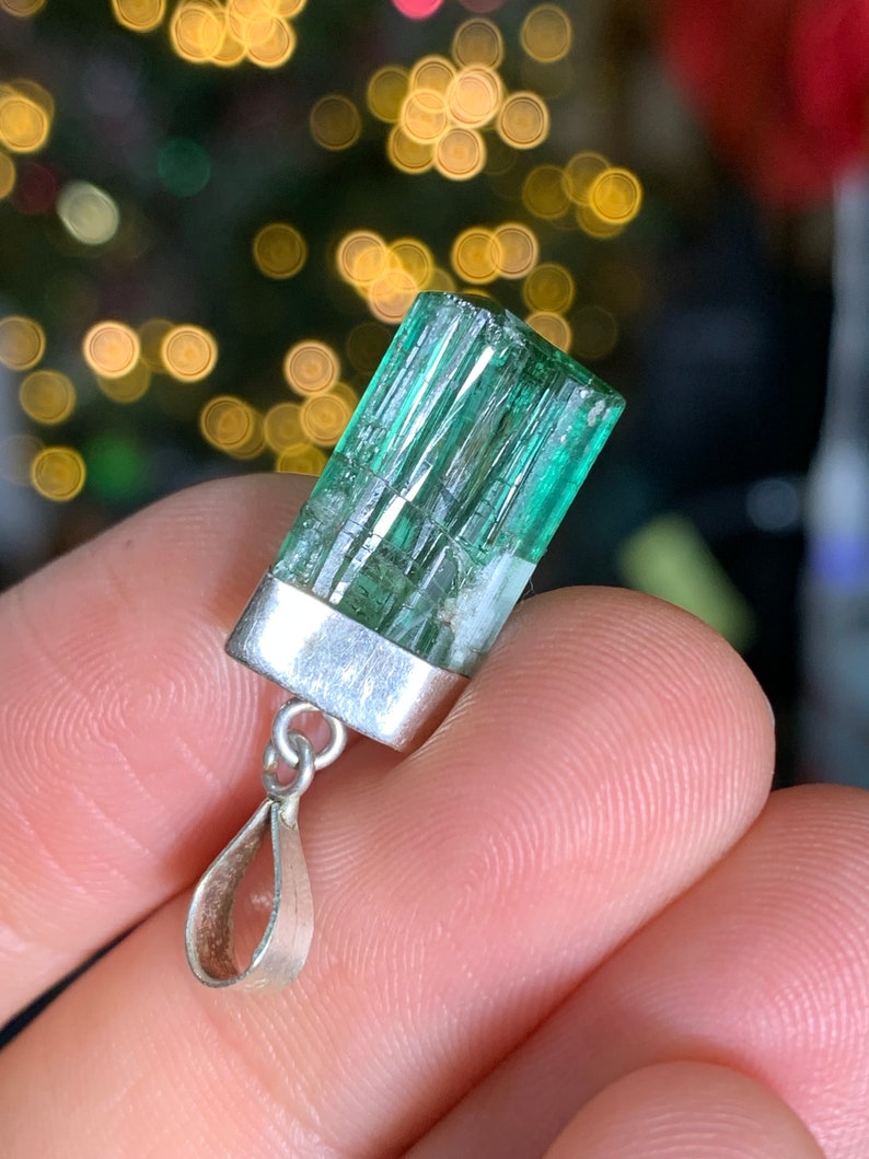 May include: A green gemstone pendant with a silver setting. The gemstone is a rectangular shape with a striated texture. The pendant is attached to a silver bail.