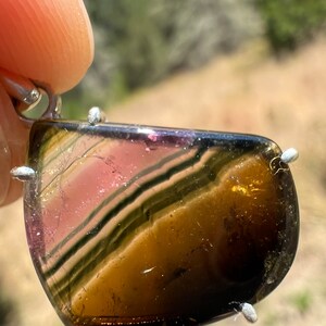 May include: A silver pendant featuring a multi-colored gemstone with brown, green, and pink stripes. The gemstone is set in a silver bezel and has a smooth, polished finish.