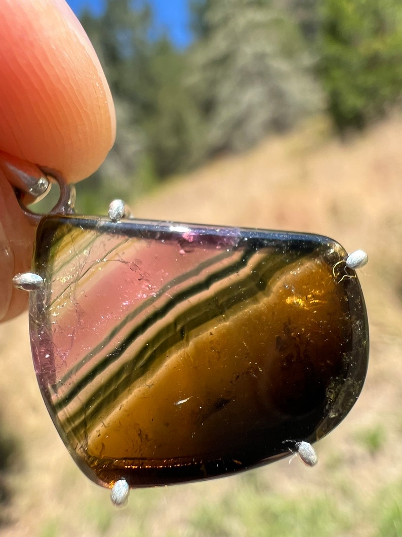 May include: A silver pendant featuring a multi-colored gemstone with brown, green, and pink stripes. The gemstone is set in a silver bezel and has a smooth, polished finish.