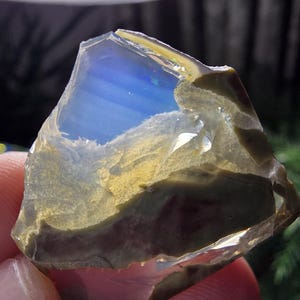 May include: A rough-cut gemstone with a translucent, layered appearance. The stone features a vibrant blue interior, contrasted by earthy tones of brown, tan, and green on the exterior. The edges are jagged, revealing the natural, raw form of the mineral.