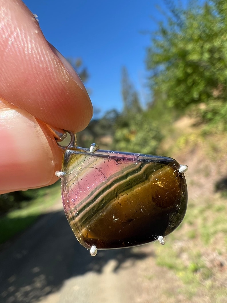 May include: A silver pendant featuring a multi-colored gemstone with green, brown, pink, and gold hues. The gemstone is set in a silver bezel with four prongs.
