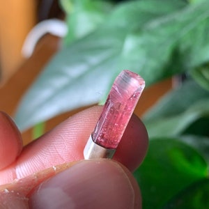 Op de afbeelding: Een kleine, langwerpige, doorschijnende roze toermalijnkristallen hanger gezet in een eenvoudige zilverkleurige metalen kap. De edelsteen heeft natuurlijke verticale strepen en een gefacetteerde bovenrand, met een levendige magentakleur.