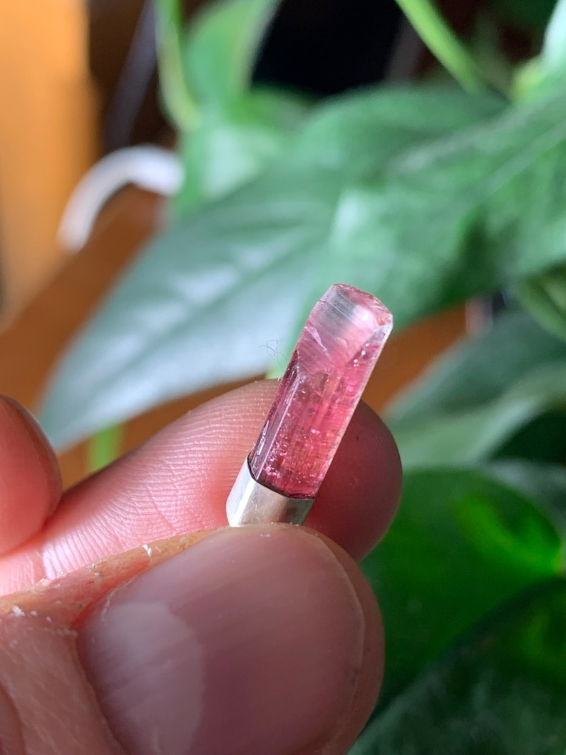 Op de afbeelding: Een kleine, langwerpige, doorschijnende roze toermalijnkristallen hanger gezet in een eenvoudige zilverkleurige metalen kap. De edelsteen heeft natuurlijke verticale strepen en een gefacetteerde bovenrand, met een levendige magentakleur.