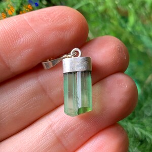May include: A raw, translucent green tourmaline crystal pendant set in a simple silver-toned metal cap with a small circular bail at the top. The crystal has a vertical, columnar structure with natural striations and a light to dark green gradient.
