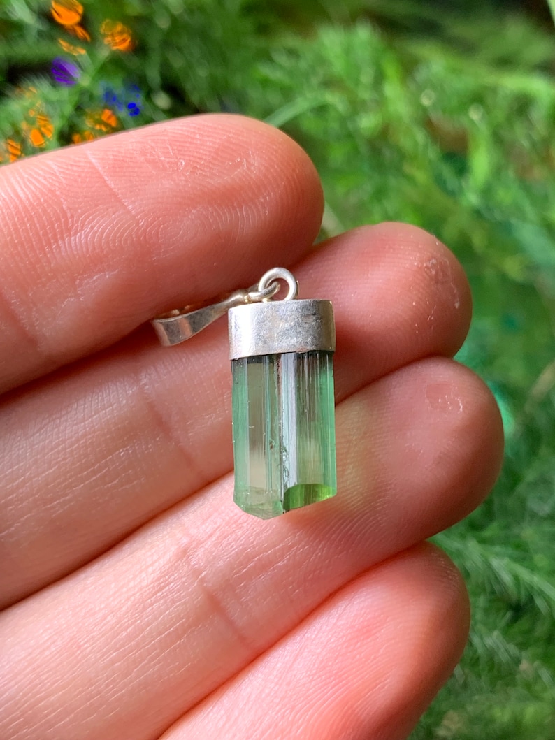 May include: A raw, translucent green tourmaline crystal pendant set in a simple silver-toned metal cap with a small circular bail at the top. The crystal has a vertical, columnar structure with natural striations and a light to dark green gradient.