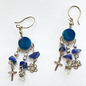 May include: A pair of dangle earrings with blue and silver beads, a blue circle, and a silver cross charm. The earrings are made of metal and have a bohemian style.