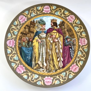 May include: A decorative ceramic plate with a detailed illustration of a king and queen in medieval attire. The plate features a floral border with pink and green accents.